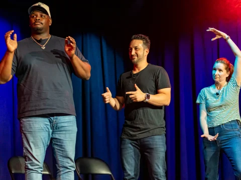 Three people performing on stage, one man in a dark T-shirt and cap gesturing, another man in a black T-shirt smiling and pointing, and a woman in a striped shirt posing with her hands up.