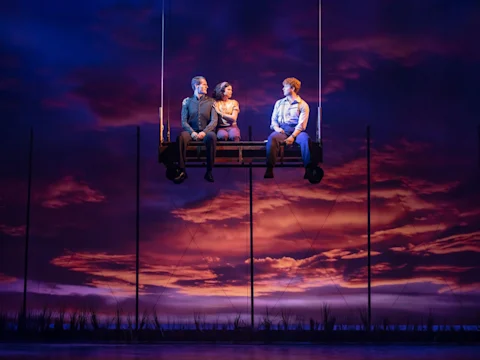 Three people sit on a suspended platform against a dramatic sunset backdrop, with stage lighting and silhouettes of tall grass below.