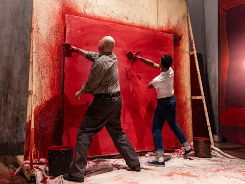 Two people paint a large canvas red with wide brushes, splattering paint onto the wall and floor around them.