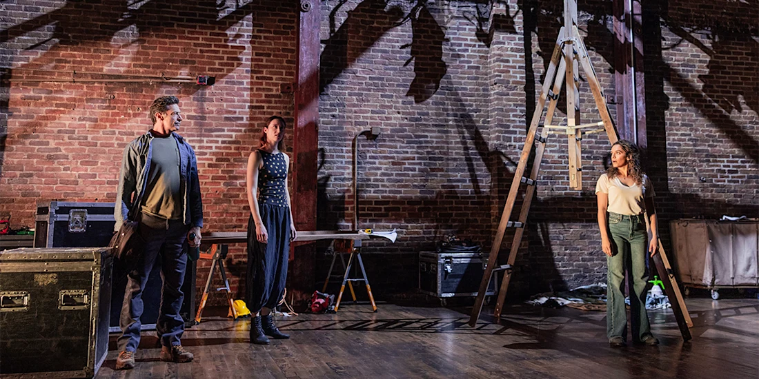 Three people stand on a stage with brick walls, scattered props, and a tall ladder, illuminated by stage lights.