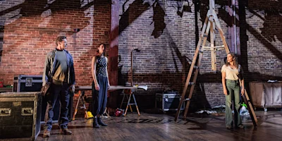 Three people stand on a stage with brick walls, scattered props, and a tall ladder, illuminated by stage lights.
