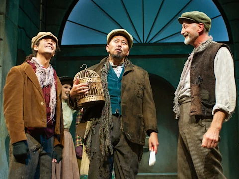 Three men in period costumes stand on stage, one holding a birdcage and another a white feather, with an arched window structure in the background.