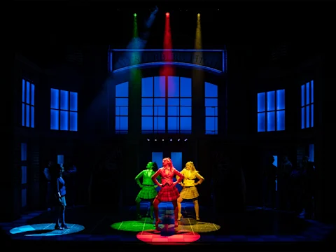 Four women in colorful costumes stand together on stage; one woman in a red outfit frames another woman's face with her hands, while the others pose beside them.