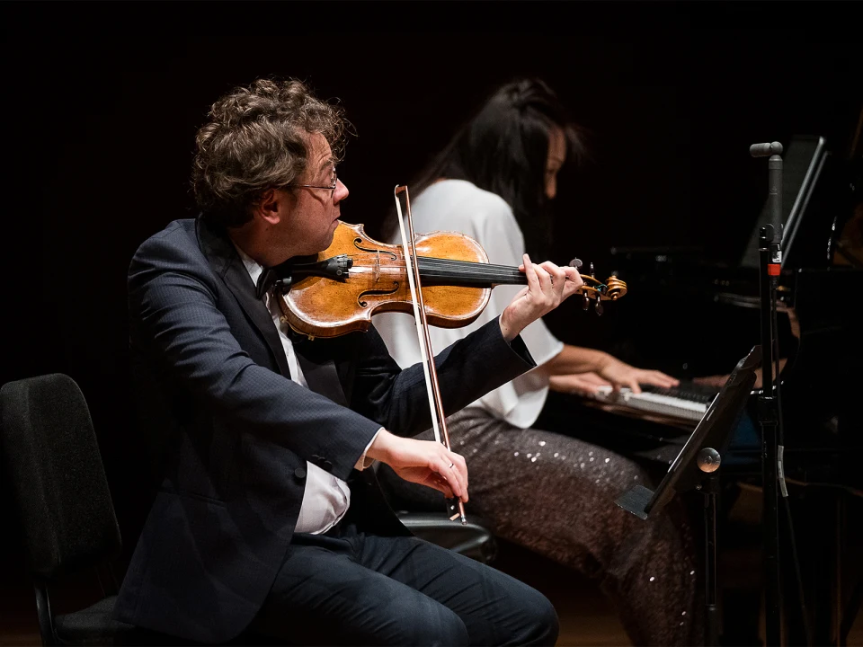 A man in a suit is playing the violin onstage, while a woman in the background plays the piano. Both are focused on their performance.