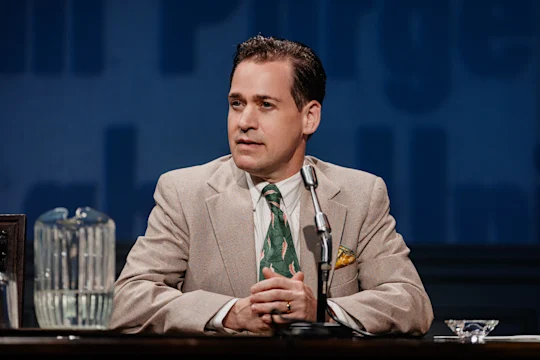 A man in a beige suit sits at a desk with a microphone, glass pitcher, and ashtray, speaking or preparing to speak in front of a blue background with partial text.
