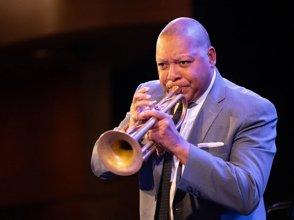 Person in a suit playing a trumpet on stage against a dark background.