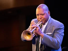 Person in a suit playing a trumpet on stage against a dark background.