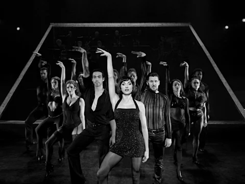 A group of performers in black outfits strike dramatic poses on stage, with a geometric backdrop and musicians visible in the background.
