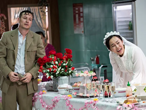 A man in a tan suit and a woman in a white bridal dress stand at a decorated table with food, drinks, and flowers in a kitchen setting. The woman is leaning and laughing.