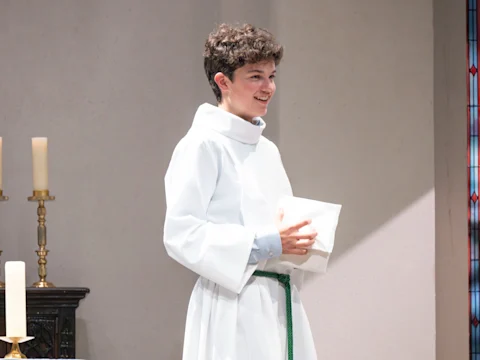 A person in a white robe with a green belt stands indoors, holding a folded white cloth. Candles and a stained glass window are visible in the background.
