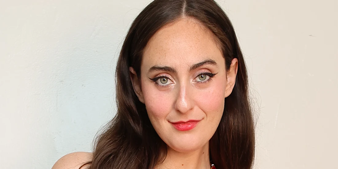 A woman with long brown hair and green eyes looks at the camera, wearing red lipstick and a sleeveless top, against a plain light-colored background.