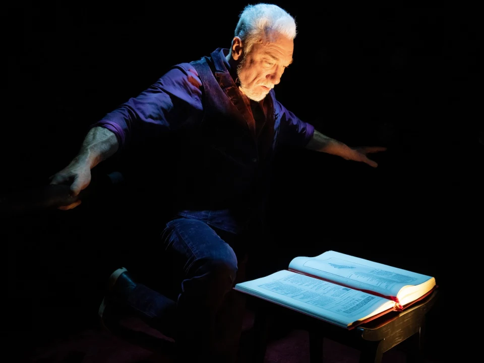 Patrick Page reading a large book with his arms stretched out to either side, in purple clothes with black rubber gloves. 