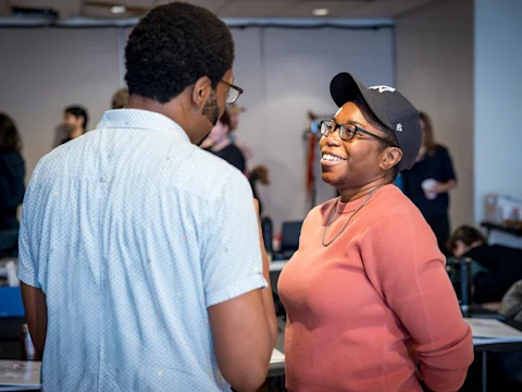 Two people stand facing each other, engaged in conversation in a room with other people in the background. One person is smiling and wearing a hat and glasses.
