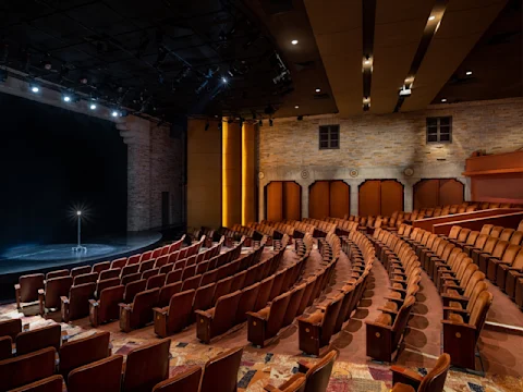 Empty theater auditorium with curved rows of brown seats facing a stage, lit by overhead lights, and stone walls in the background.