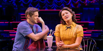 Two people sit at a table with a milkshake and two straws, talking under stage lights with a colorful, theatrical backdrop.