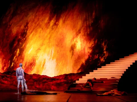 A dramatic stage scene with a fiery backdrop, a nude figure standing, three people lying on the ground, and a red staircase at the right.