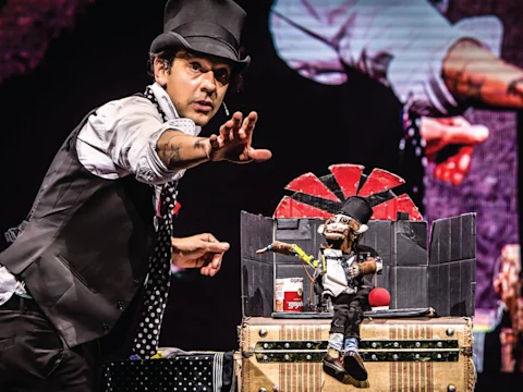 A magician in a top hat and vest performs an act with a small puppet on a stage, using props including a red-and-black spinning wheel and a can.