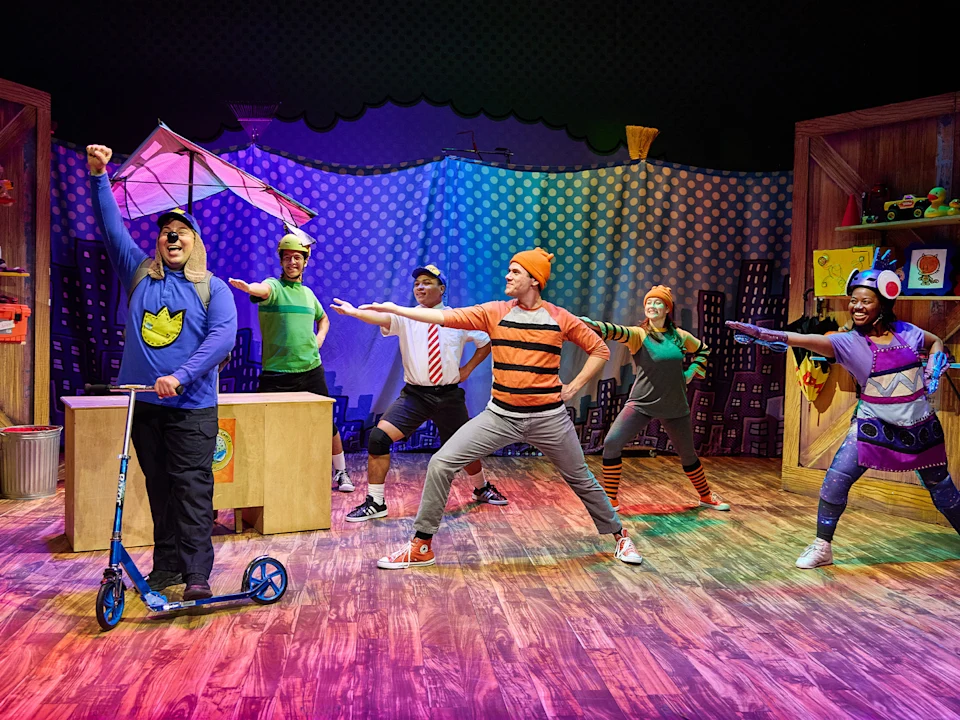 Five performers in colorful costumes pose energetically on a stage with playful props, a scooter, and vibrant lighting, suggesting a children's theater production.