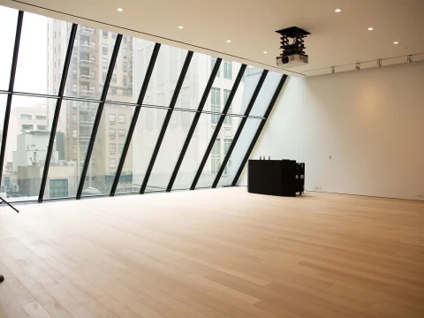 An empty room with wooden flooring, angled glass wall, ceiling lights, and a black podium. Urban buildings are visible through the glass.