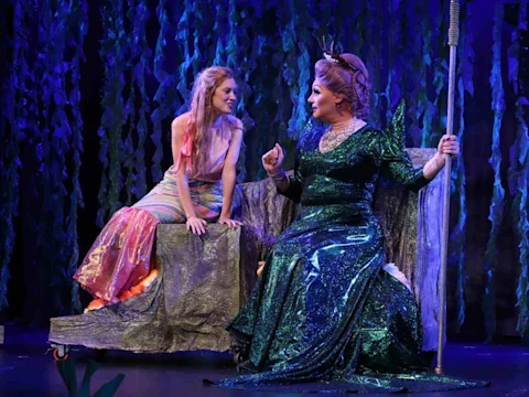 Two performers in colorful costumes sit on stage, one in a shimmery mermaid outfit and the other in a dark green gown holding a trident, against a backdrop of foliage.