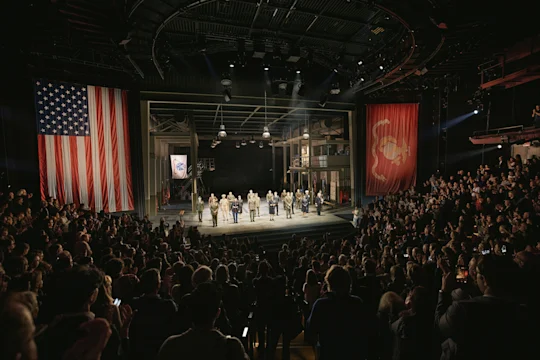 A group of military personnel in formal uniforms stand on a stage before an audience in a theater, flanked by large American and Marine Corps flags.
