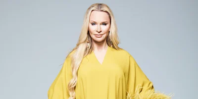 A woman with long blonde hair wears a loose-fitting yellow dress and large earrings, standing against a plain light gray background.