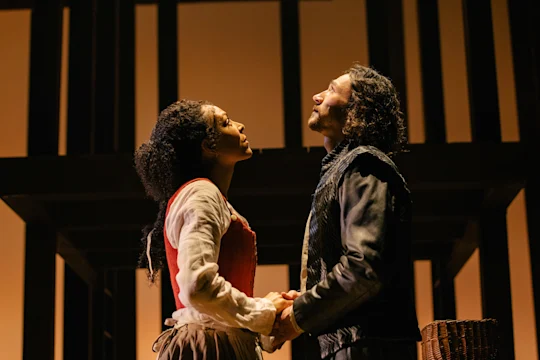 Two actors in period costumes stand close together on stage, holding hands and looking upward against a backdrop of vertical and horizontal beams.