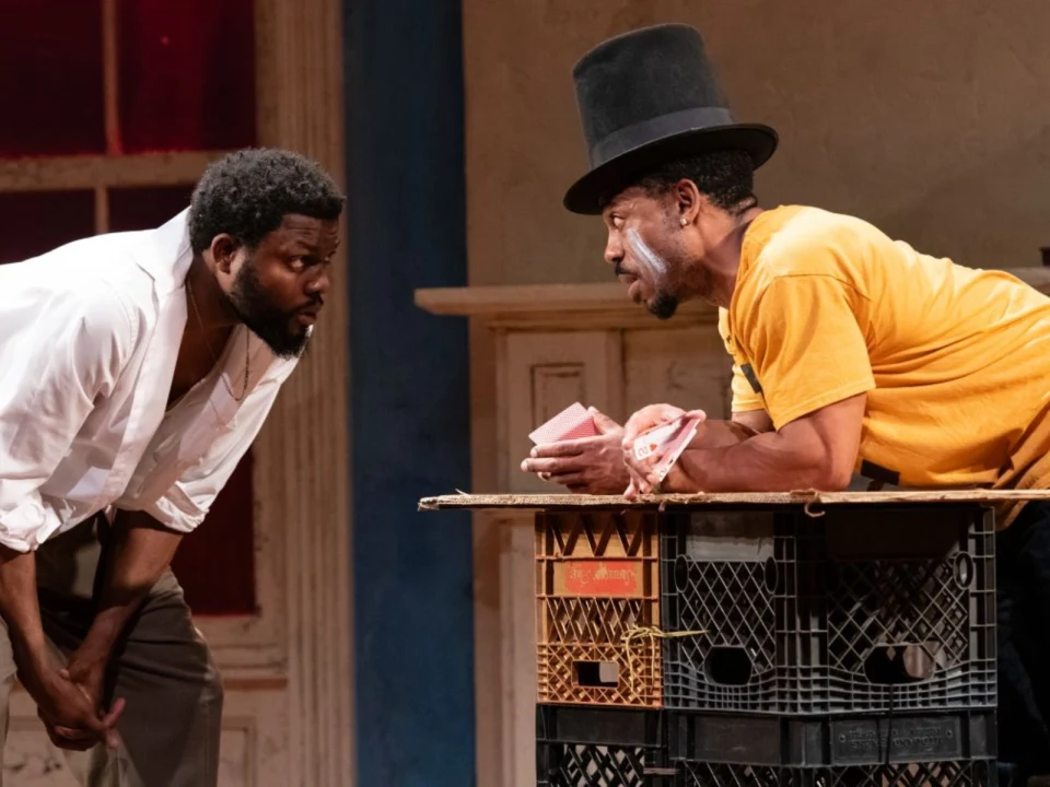 Two men engage in an intense discussion in a dimly lit room. One man leans forward wearing a white shirt, while the other, in a yellow shirt and top hat, holds a stack of cards over a makeshift table.