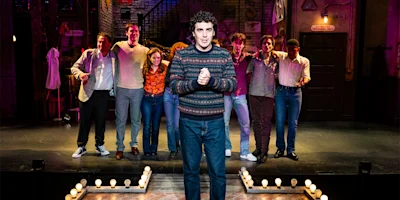 A man in a patterned sweater stands at the front of a stage, with seven people standing side by side in the background under stage lights.