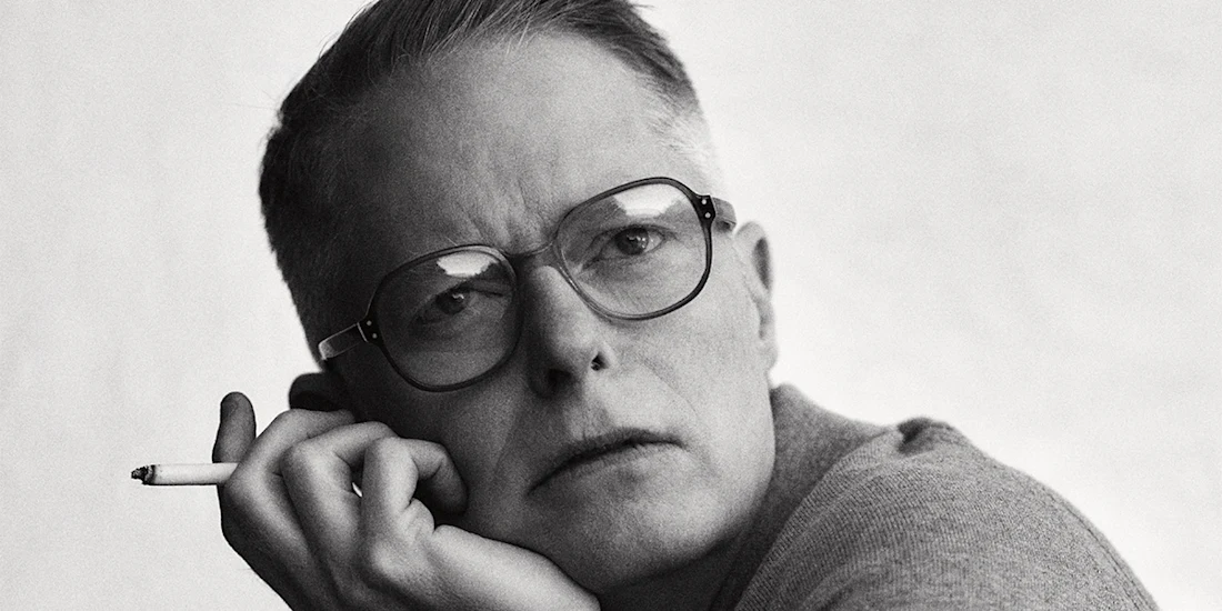 Person wearing glasses and a sweater holds a cigarette, resting their head on their hand, and looks thoughtfully towards the camera.