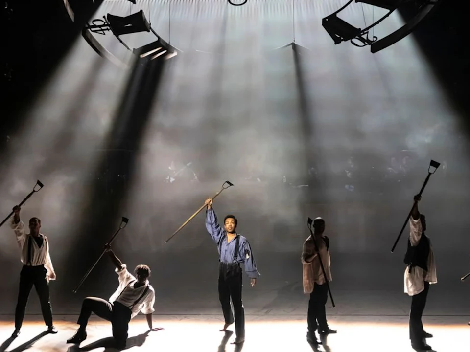 Five performers on stage hold axes aloft under dramatic overhead lighting, with light and shadows emphasizing their poses.