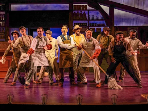 A group of actors dressed as workers perform energetically on stage, holding mops and shovels, with kitchen shelves in the background.