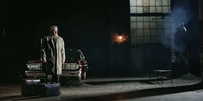 A man in a trench coat stands beside two suitcases in a dimly lit garage, with an old red car and industrial windows in the background.