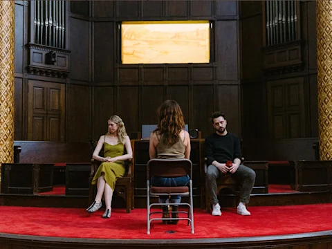 Three people sit on stage in a wood-paneled room with red carpet; one faces away from the camera, while two others sit apart facing forward.