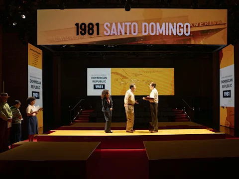 Five people stand on a stage set with large screens displaying “1981 SANTO DOMINGO” and “Dominican Republic 1981.” Two people in the center appear to be conversing.