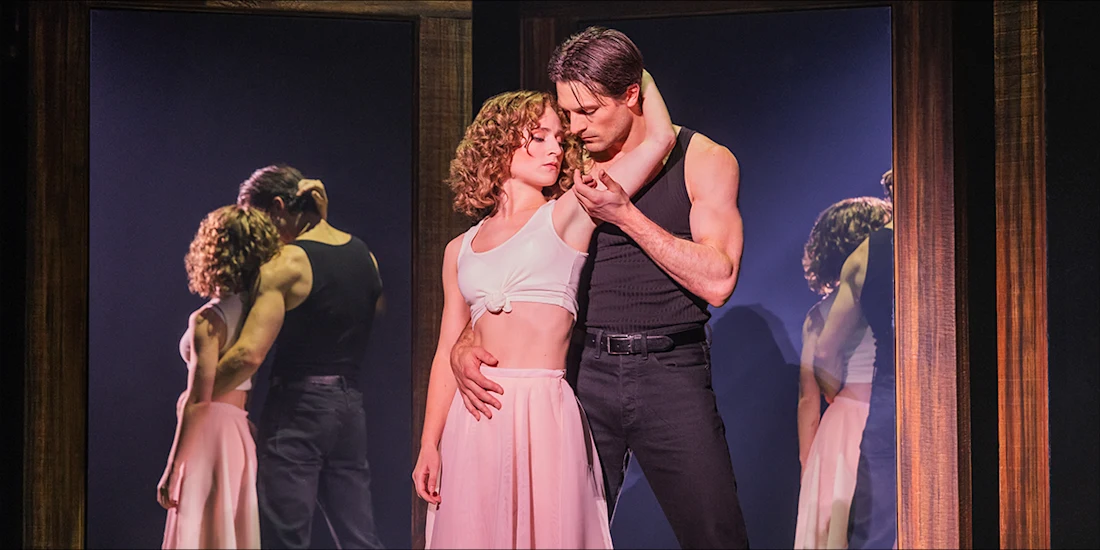 A man and a woman in dance attire embrace in front of two large mirrors.