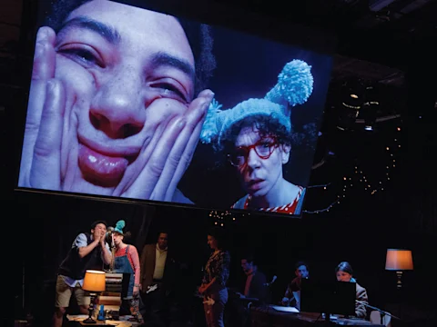 Large screen displays two people making faces; one squishes cheeks and the other wears a blue hat. Below, a group of people are engaged in various activities on stage with dim lighting.