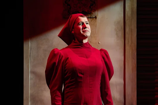 A person in a red, long-sleeved dress and head covering stands under a beam of light against a textured wall.