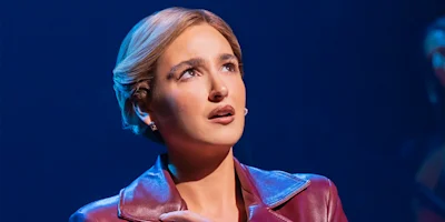 A person with short hair and a concerned expression wears a maroon jacket, standing against a dark blue background under stage lighting.