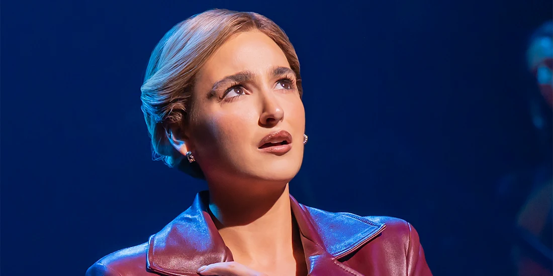 A person with short hair and a concerned expression wears a maroon jacket, standing against a dark blue background under stage lighting.