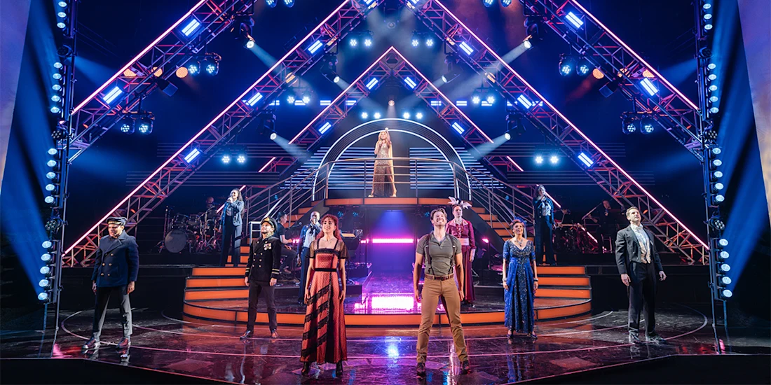 A group of performers stand on a brightly lit stage with geometric lighting structures and staircases, while a singer stands elevated at the center.