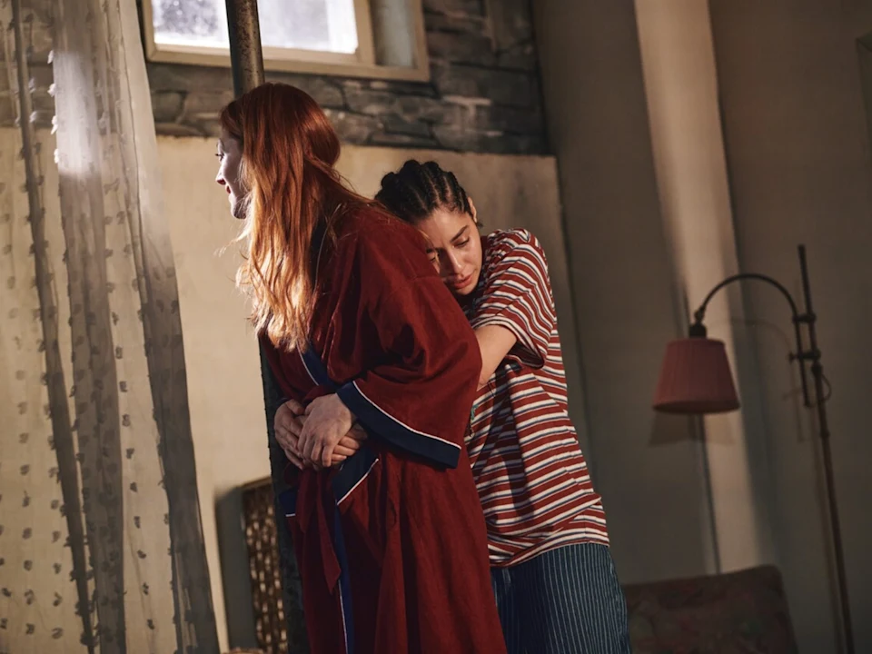 Two women stand indoors; one in a red robe faces away, while the other in a striped shirt hugs her from behind, both appearing thoughtful.