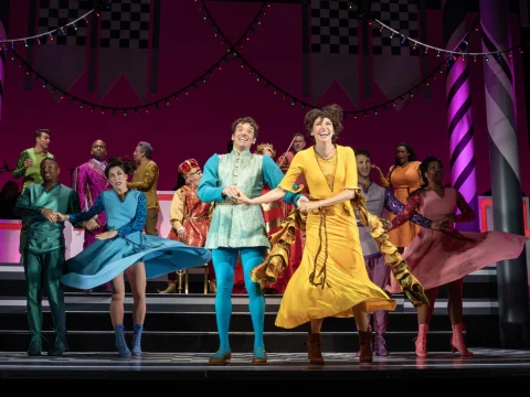 A group of people in colorful costumes perform a lively dance on stage, with vibrant lighting and decorated background elements suggesting a theatrical production.