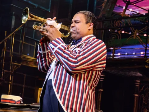 A person in a striped jacket plays a trumpet on stage with colorful lights and a musical setting in the background.