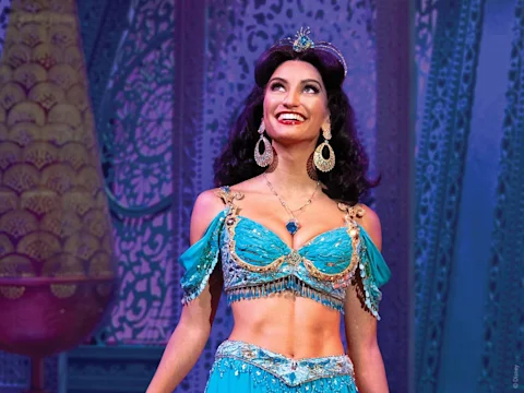A woman in a blue, jeweled costume with gold accents smiles on stage, standing in front of an ornate, patterned backdrop.