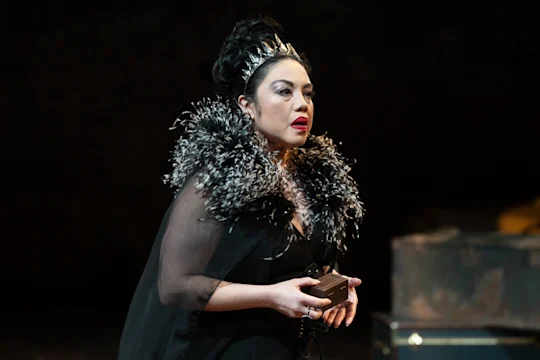 A person in a dramatic black costume with a feathered collar and a crown holds an item on stage.