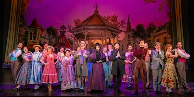 A large cast in colorful period costumes stands on stage in front of a gazebo, with a painted backdrop of a quaint town and pink sky, likely from a musical or theatrical performance.