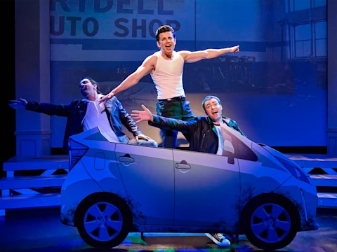 Three men in costumes pose energetically inside a painted car prop on stage, with a backdrop reading “Rydell Auto Shop” behind them.
