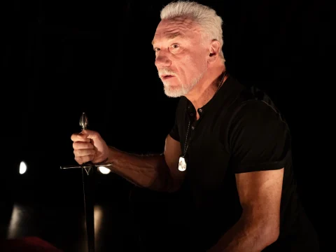 Patrick Page holding a sword in a black t shirt looking concerned.