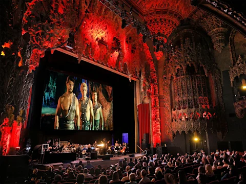 An audience watches a movie screening with live music in an ornate, red-lit theater featuring elaborate ceiling and wall carvings.